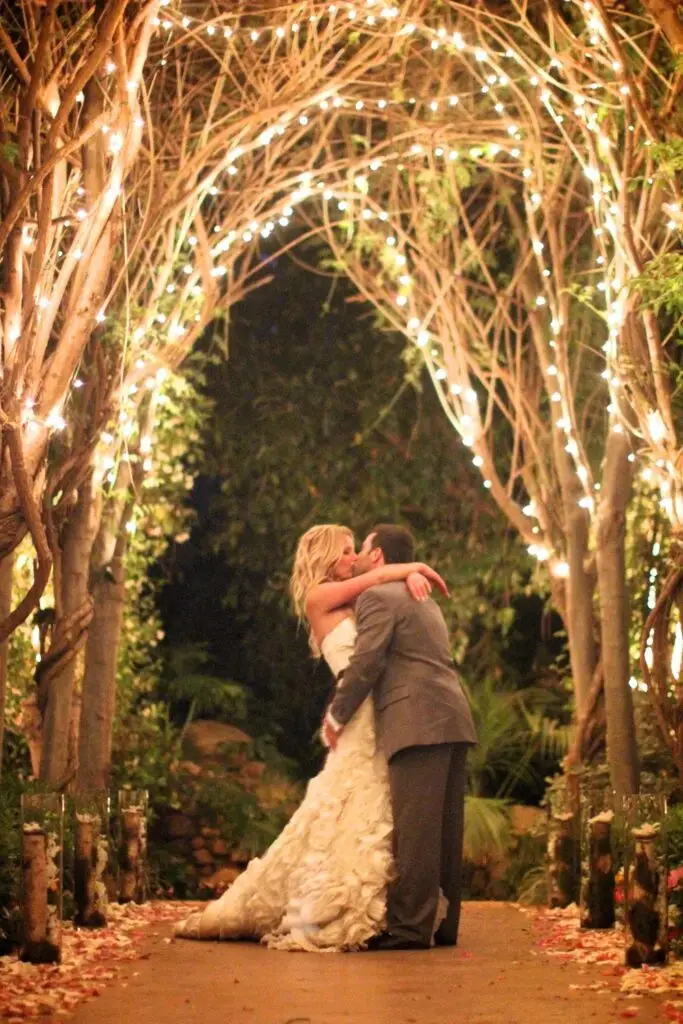 Couple kissing under lighted tree arch at wedding.